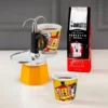 Bialetti Mini Express Lichtenstein Moka Pot with one cup on platform on on side filled with coffee and a bag of Bialetti Perfetto Moka coffee