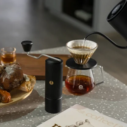Chestnut X-lite Manual Portable Coffee Grinder on a table with pastries and a pour over being poured from a Fish pour over kettle